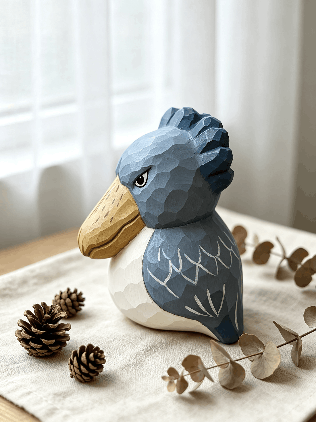A handcrafted low-poly wooden shoebill figurine in slate blue and cream, arranged on linen cloth with pine cones and dried eucalyptus in natural window light