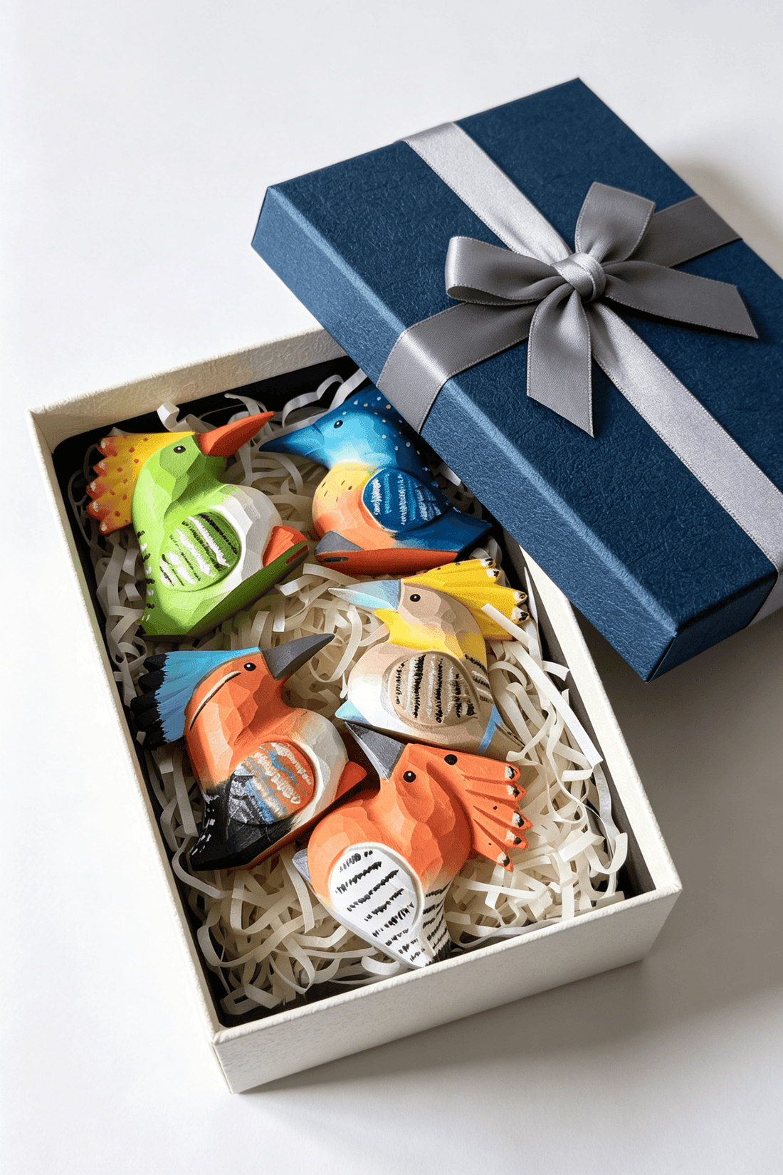 A set of five colourful carved wooden bird figurines arranged inside a dark blue gift box lined with shredded paper