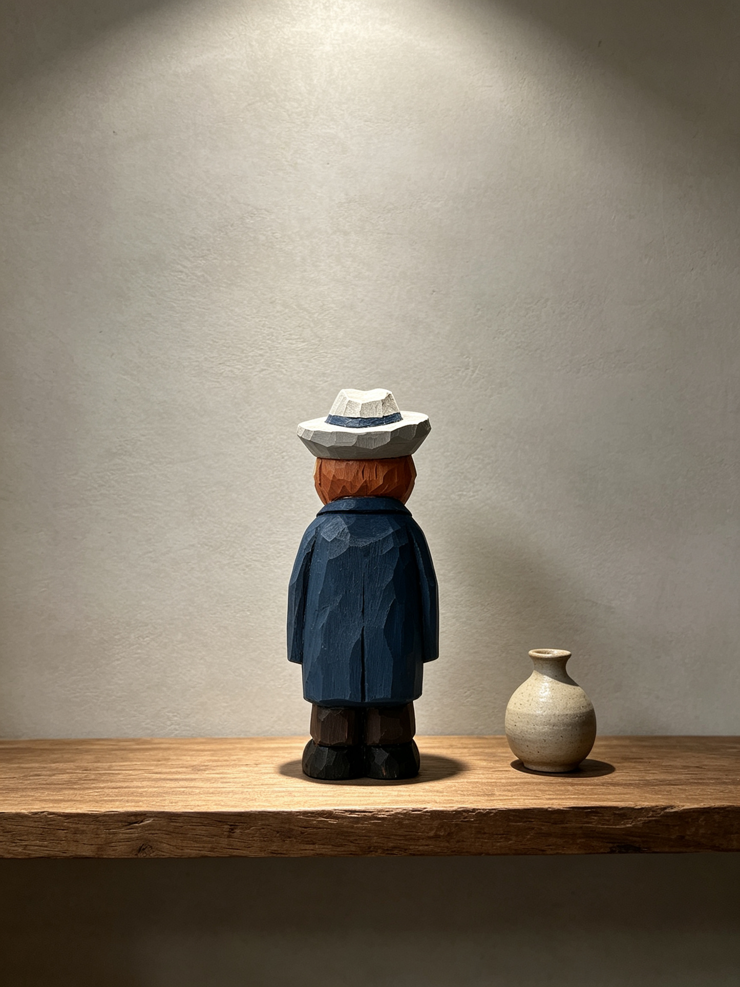 A low-poly wooden figurine of a person seen from the back, wearing a white hat, reddish-brown hair, a blue coat, and dark pants, standing on a wooden shelf next to a small beige ceramic vase against a textured gray wall.