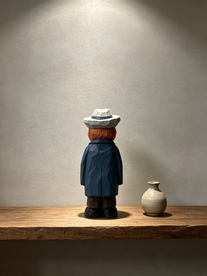 A low-poly wooden figurine of a person seen from the back, wearing a white hat, reddish-brown hair, a blue coat, and dark pants, standing on a wooden shelf next to a small beige ceramic vase against a textured gray wall.