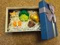 A cream-colored gift box with a blue lid and silver ribbon, containing hand-carved low-poly figurines of two orange corgis, a cute green broccoli character, and a golden cloud-like creature resting on white shredded paper.