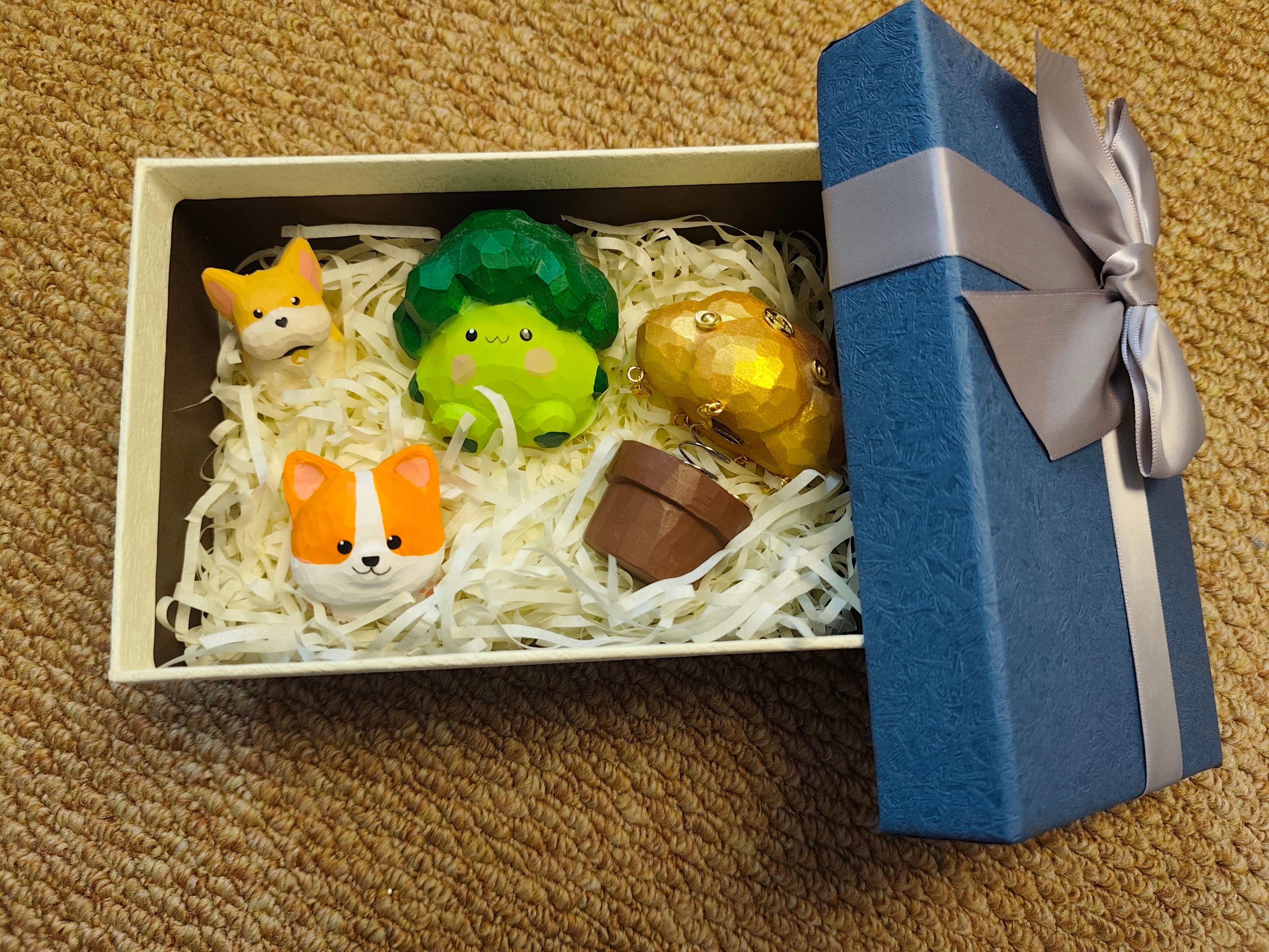 A cream-colored gift box with a blue lid and silver ribbon, containing hand-carved low-poly figurines of two orange corgis, a cute green broccoli character, and a golden cloud-like creature resting on white shredded paper.